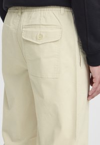 Pantaloni bej din bumbac cu buzunar posterior cu nasture și talie elastică, prezentați din profil. Textură netedă și croială lejeră.