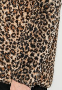 Rukáv huňatej bundy s leopardím vzorom v odtieňoch hnedej, béžovej a čiernej farby, pokrývajúci ruku osoby a zhotovený z mäkkej textúrovanej látky.
