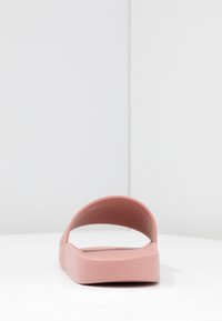 Růžové gumové šlapky se zakřiveným páskem, texturovaným povrchem a plochou podrážkou. Jednoduchý design, minimální detaily a žádné kovové prvky.