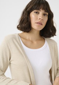 Beige lange cardigan over een witte ribbel-tanktop, met een open voorkant ontwerp en een zachte textuur, geschikt voor het lagen.