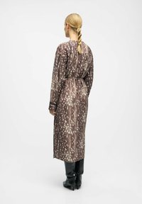 Femme aux cheveux blonds tressés portant une longue robe marron à taches blanches, bottes noires à talons, debout de dos sur un fond uni.