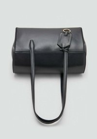 Bolso de piel negra con forma cilíndrica, con un asa larga y una etiqueta, textura lisa y diseño minimalista.