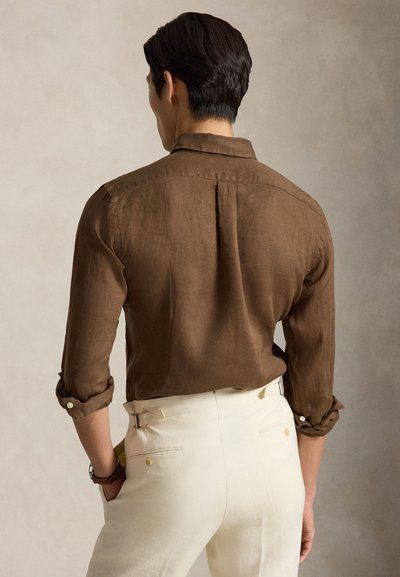 Bărbat cu păr închis la culoare, purtând o cămașă maro cu mâneci lungi și manșete suflecate și pantaloni de culoare crem, stând cu mâinile în buzunare, vedere din spate.