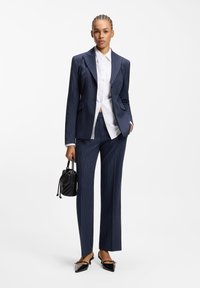Costume pantalon rayé marine avec un blazer ajusté, une chemise blanche à boutons, des chaussures noires pointues et un petit sac à main noir.