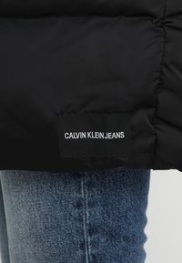 Musta toppatakki mattaisen pinnan kanssa, jonka päällä näkyy selkeä valkoinen etiketti, jossa lukee "CALVIN KLEIN JEANS", ja alla siniset farkut.