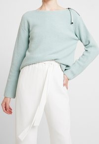 Hellblaues, geripptes Strickoberteil mit weitem Ausschnitt und Reißverschlussdetail an der Schulter, kombiniert mit weißen, hochgeschnittenen Hosen.