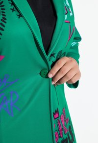 Een groene knoop vastmaken op een blazer met graffiti-print, gedragen over een zwart topje, cropped tot aan de torso met een witte achtergrond.