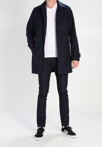 Manteau en mélange de laine bleu marine avec col cranté, fermeture à boutons et deux poches avant, associé à un jean foncé et des baskets noires.