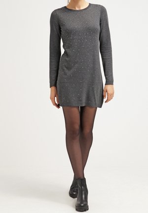 Kobieta ubrana w szarą, dopasowaną mini sukienkę z długim rękawem i małymi białymi kropkami, czarne przezroczyste rajstopy oraz czarne botki, stojąca na jednolitym tle.