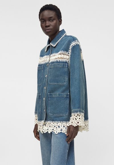 Γυναίκα που φοράει μπλε τζιν μπουφάν και τζιν, με λευκή δαντέλα κροσέ στο γιακά, στο στήθος, στα μανίκια και στο κάτω μέρος.