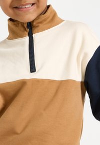 Mosolygó gyermek, színblokkolt pulóverben, amelynek barna gallérja, krémszínű melle, barna törzse és tengerészkék ujja van, fekete cipzárral.