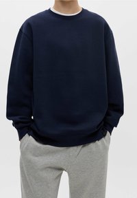 Felpa blu navy con scollatura rotonda, spalle scese e polsini a costine. Abbinata a pantaloni della tuta grigi chiari, caratterizzati da una vestibilità comoda.