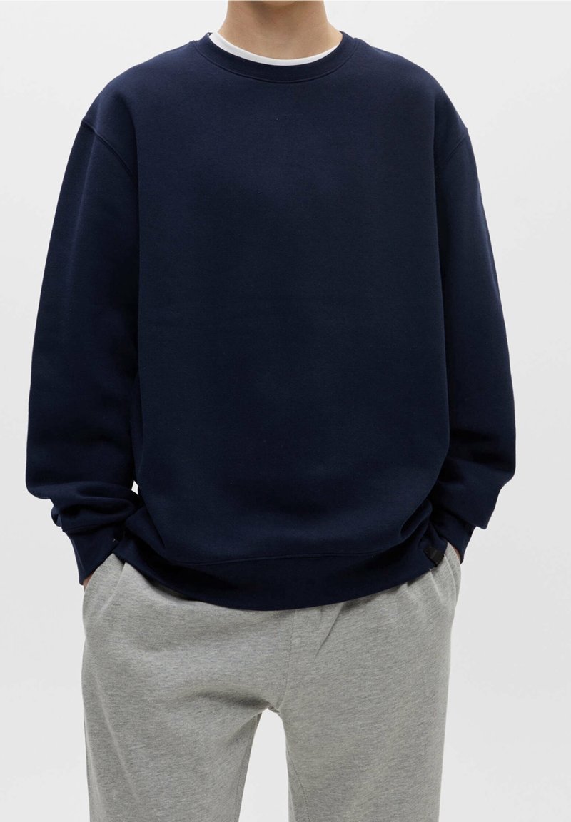 Felpa blu navy con scollatura rotonda, spalle scese e polsini a costine. Abbinata a pantaloni della tuta grigi chiari, caratterizzati da una vestibilità comoda.