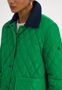 Grønn quiltet jakke med marineblå krage, frontlommer og en logo på brystet. Knapper synlige langs knapperaden. Glatt tekstur.