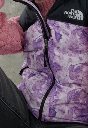 Henkilö, jolla on päällään violetti kuvioitu North Face -takki, pinkki neulottu käsine ja harmaat housut harmaata betonitaustaa vasten.
