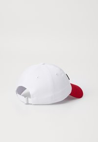 Casquette de baseball blanche avec un pic rouge, présentant une forme courbée, une sangle ajustable et un accent noir sur le côté. Texture en tissu coton.