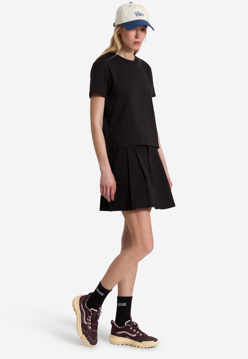 Femme portant un t-shirt noir et une jupe plissée, une casquette beige et bleue, des chaussettes noires Vans et des baskets violet foncé avec des semelles beiges, debout de profil.