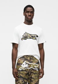 Άσπρο βαμβακερό t-shirt με μαύρο και καμουφλάζ γραφικό σχέδιο. Συνδυασμένο με παντελόνι καμουφλάζ cargo, που παρουσιάζει λεπτομέρεια με λευκή βαφή σπρέι.