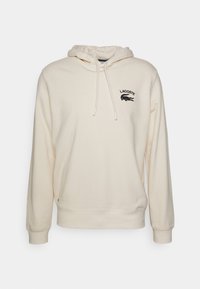 Sweat à capuche de couleur crème en coton doux, avec une capuche à cordon, des poignets côtelés et un logo crocodile Lacoste noir sur la poitrine.