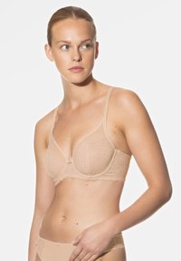 Soutien-gorge en dentelle beige avec armatures, motif floral, bords festonnés et un petit nœud au centre. Bretelles ajustables et texture douce.