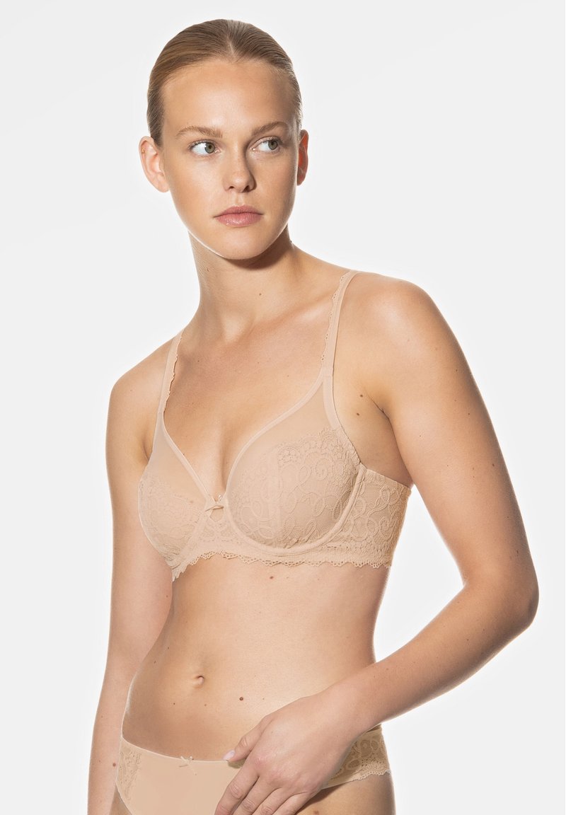 Soutien-gorge en dentelle beige avec armatures, motif floral, bords festonnés et un petit nœud au centre. Bretelles ajustables et texture douce.