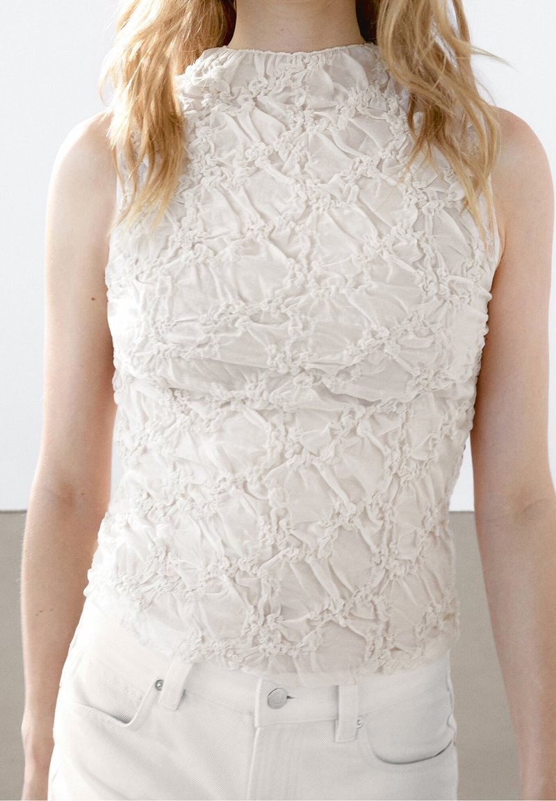 Femme portant un haut blanc sans manches à col montant avec un motif floral texturé froncé, associé à un pantalon blanc.