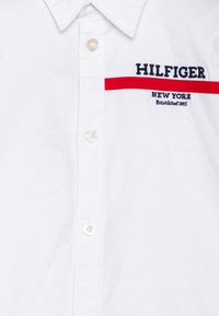 Bela bombažna srajca s kragnim izrezom, ki vsebuje mornarsko plavo vezeno besedilo "HILFIGER" ter rdečo črto z "NEW YORK" in "Ustanovljeno 1985."