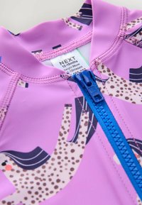 Maillot de bain pour enfants rose avec une fermeture éclair bleue. Présente des impressions d'animaux en crème et en couleurs foncées. Tissu lisse et extensible. Étiquette de taille visible.