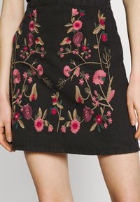 Svart kjol med rosa, röd och beige blommotiv broderi, bärs av en person med händerna vilande vid sidan av kjolen.