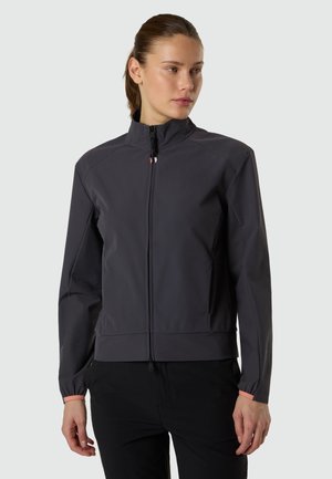 Donna con giacca grigia scura con zip e colletto alto, pantaloni neri, che guarda leggermente verso destra su uno sfondo neutro.