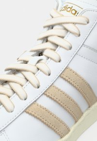 Λευκό δερμάτινο αθλητικό παπούτσι με ρίγες από μπεζ σουέτ, με μπεζ κορδόνια και υφασμένη σόλα. Χρυσό λογότυπο "adidas" κοντά στη γλώσσα.