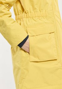 Veste extérieure jaune avec une taille cintrée, dotée d'une poche avant et d'une texture lisse. La manche a une doublure intérieure noire.