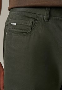 Chinos vert olive avec une coupe slim, dotés d'une poche arrière et de détails en métal argenté. Tissu texturé avec une finition lisse.