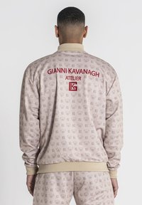 Lichtbeige jasje met herhalend logo-patroon, geribde manchetten en zoom, en rood geborduurde tekst "GIANNI KAVANAGH ATELIER" op de achterkant.