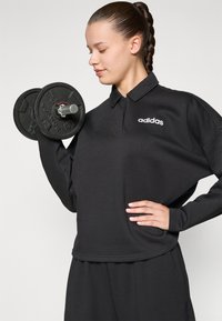 Melnas, garām piedurknēm polo krekls ar mīkstu tekstūru un īsu apakšmalu. Iespējots ar baltu adidas logo un papildināts ar hanteli.
