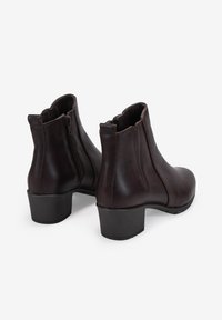Botas de tobillo de cuero marrón con un tacón bajo en bloque, punta redondeada y cierre de cremallera lateral con bordes ondulados en la parte superior.