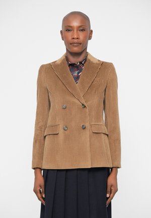 Woman wearing a tan corduroy double-breasted blazer over a patterned shirt and dark pleated skirt, standing against a plain white background.