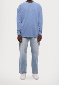 Camicia azzurra a maniche lunghe con logo ricamato sul petto, abbinata a jeans azzurri larghi con dettagli sfilacciati e sneakers bianche.