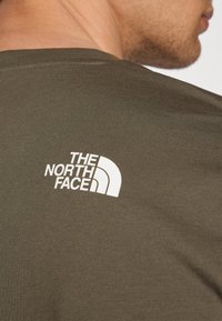 Oliivinvihreä t-paita pehmeästä kankaasta, jossa on näkyvä valkoinen logo The North Face -merkin yläselässä. Yksinkertainen pyöreä pääntie.