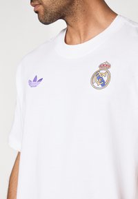 T-shirt branca de algodão com um logótipo Adidas roxo e um emblema do Real Madrid bordado e multicolorido no lado esquerdo do peito. Mangas curtas.