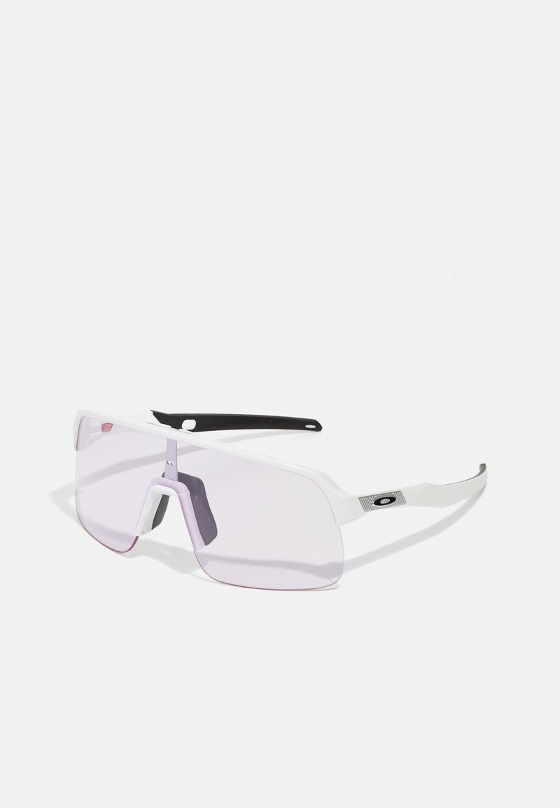Gafas de sol grandes y blancas con lentes tintados de púrpura, un diseño de montura prominente y acentos negros en las patillas. Presenta un puente frontal distintivo.