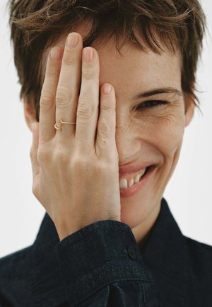 Persona sonriente con el cabello corto que se cubre un ojo con la mano, lleva un anillo de oro y viste una camisa oscura de manga larga.