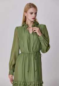 Femme portant une robe longue à manches longues olive avec des détails de col fleuri, une taille cintrée à nouer, et un ourlet à volants, regardant sur le côté devant un fond uni.