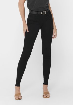 Femme portant un pantalon skinny noir avec une ceinture noire et des sandales ouvertes couleur beige, debout devant un fond uni.