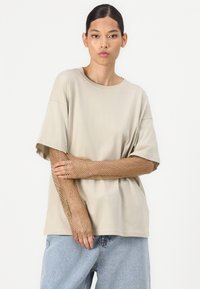 Beige oversized T-shirt met korte mouwen, gecombineerd met lange, metalen nethandschoenen. Model poseert met gekruiste armen en toont textuurdetails.