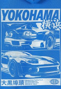 Modra grafična majica s kratkimi rokavi z dvema športnima avtomobiloma, obrisanima z belo barvo. Napis vsebuje "YOKOHAMA" in druge lokacijske podatke na dnu.