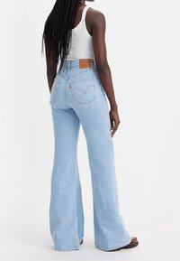 Kvinna med långt flätat hår som bär ljusblå högmidjade utsvängda jeans, vit linne och bruna skor, stående mot en enfärgad vit bakgrund.