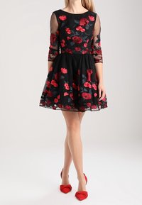 Černé šaty s tříčtvrtečními průsvitnými rukávy, vyšívanými červenými květinami, přiléhavým živůtkem, rozšířenou sukní a vlnkovaným lemem. Ukomponováno s červenými botami.