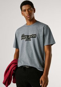 Sivé bavlnené tričko s čiernobielym logom „PEPE JEANS LONDON“, krátkymi rukávmi a voľným strihom, držené spolu s červenou mikinou s kapucňou.