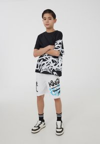 T-shirt nera con design grafico bianco su una manica, abbinata a pantaloni corti bianchi con testo graffiti nero e blu. Sneakers in bianco e nero.